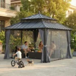 SUNJOY Kingston 12x12 gazebo with gray powder‑coated aluminum posts, galvanized steel hardtop, dual vented roof, and beige curtains/netting installed on a wooden deck.